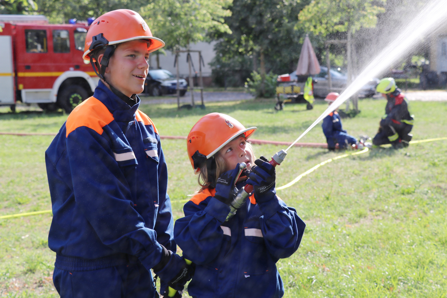 Zwei Jugendliche in Feuerwehruniform mit orangefarbenen Helmen halten gemeinsam ein spritzendes Strahlrohr; im Hintergrund ein Löschfahrzeug und weitere Übende.