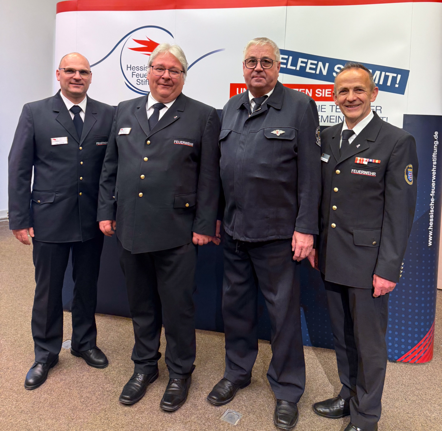 Vier Männer in dunkler Feuerwehruniform stehen nebeneinander vor einer Messewand der Hessischen Feuerwehrstiftung.