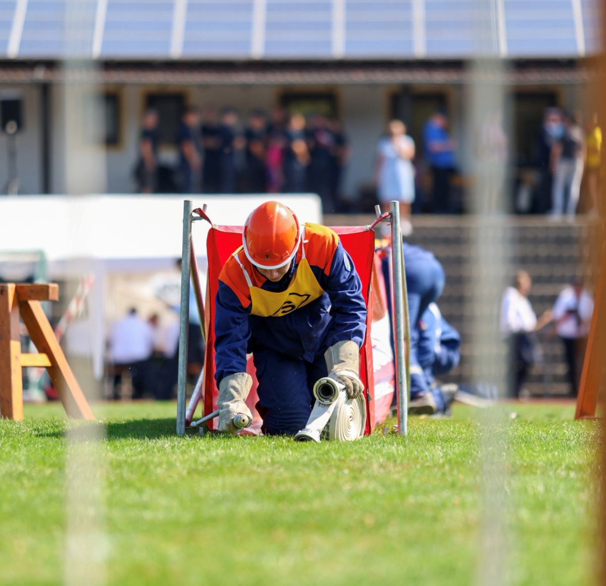 Jugendfeuerwehrmitglied mit orangefarbenem Helm rollt beim Wettbewerb auf einer Rasenfläche einen Schlauch aus; im Hintergrund Zuschauer auf einer Tribüne.