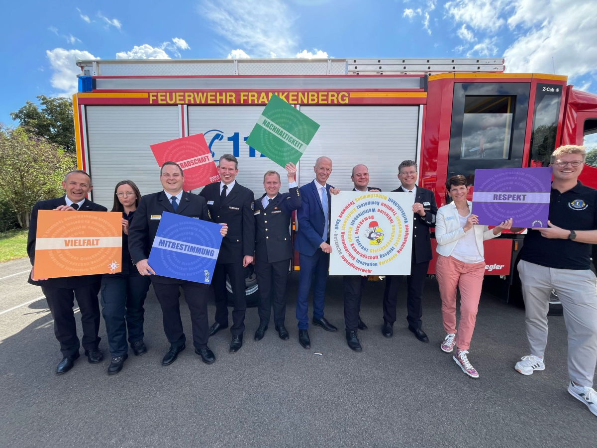 Gruppe von Personen, teils in Feuerwehruniform, steht vor einem Löschfahrzeug der Feuerwehr Frankenberg und hält farbige Schilder mit Wertebegriffen wie Vielfalt, Mitbestimmung, Nachhaltigkeit und Respekt hoch.