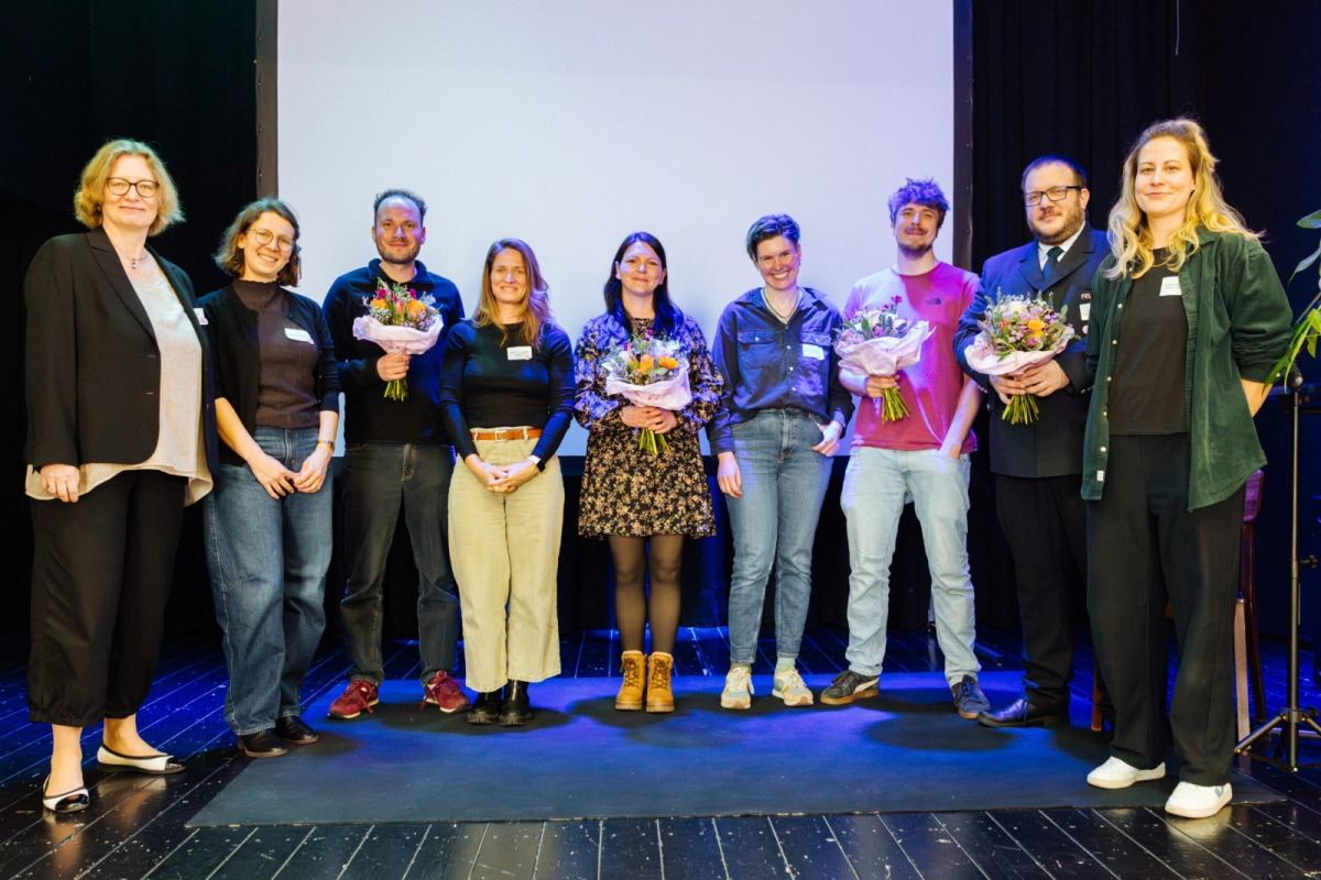 Gruppe von neun Personen auf einer Bühne, einige mit Blumensträußen, beim Vernetzungstreffen zur queeren Jugendarbeit