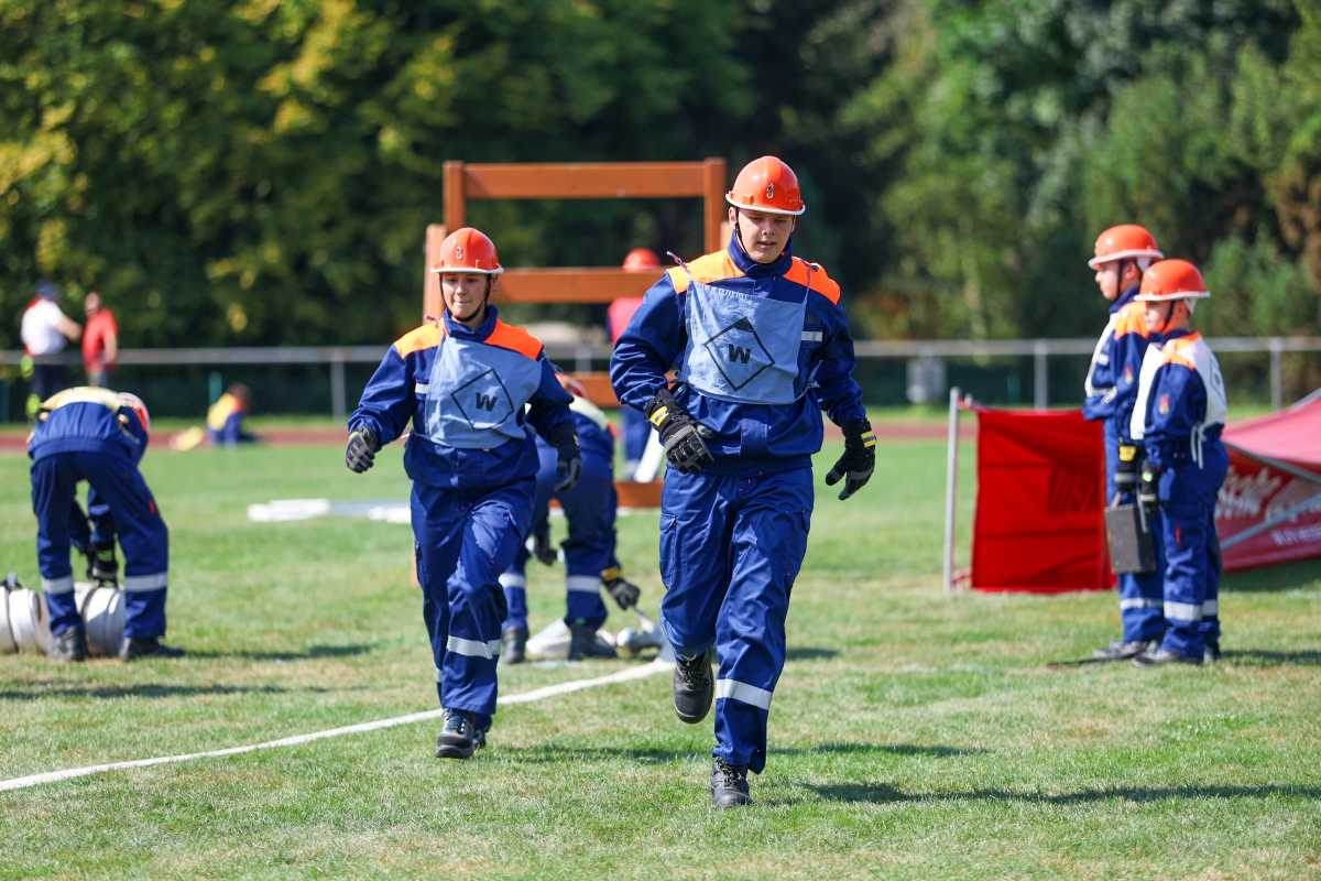 Mehrere Jugendliche in blauen Uniformen und orangefarbenen Helmen laufen über eine Wettbewerbsbahn auf einer Rasenfläche